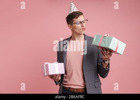 Image d'un jeune homme sérieux de pensée dans des verres tenant anniversaire cadeau boîtes isolées sur fond de mur rose. Banque D'Images