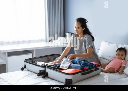 le bébé mignon se trouve à côté de la mère préparant des vêtements à mettre dans la valise Banque D'Images