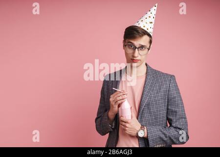 Portrait d'un jeune homme bouleversé portant un chapeau d'anniversaire célébrant isolé sur fond rose, tenant une boisson Banque D'Images