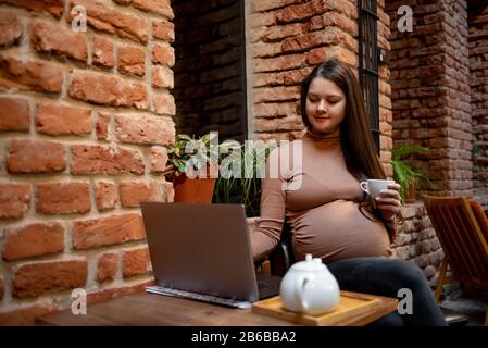 Portrait de jeune femme indépendante prégnanty utilisant un ordinateur portable pour un travail à distance tout en étant assis dans un café moderne avec une tasse de thé Banque D'Images