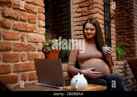 Portrait de jeune femme indépendante prégnanty utilisant un ordinateur portable pour un travail à distance tout en étant assis dans un café moderne avec une tasse de thé Banque D'Images