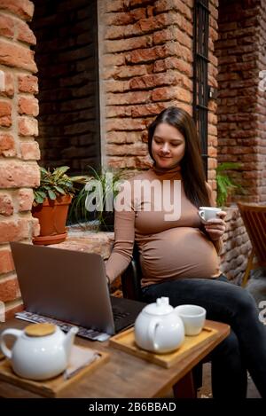 Portrait de jeune femme indépendante prégnanty utilisant un ordinateur portable pour un travail à distance tout en étant assis dans un café moderne avec une tasse de thé Banque D'Images