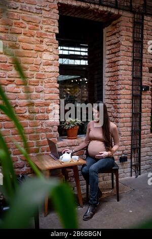 Portrait de jeune femme indépendante prégnanty utilisant un ordinateur portable pour un travail à distance tout en étant assis dans un café moderne avec une tasse de thé Banque D'Images