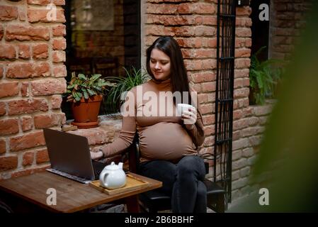 Portrait de jeune femme indépendante prégnanty utilisant un ordinateur portable pour un travail à distance tout en étant assis dans un café moderne avec une tasse de thé Banque D'Images