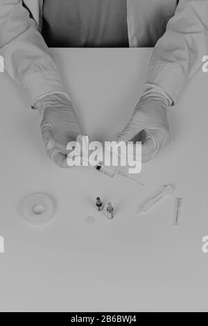 MD préparation de l'injection, style monochrome délavé. Vue de dessus des mains du médecin dans les gants et la seringue de maintien du revêtement de laboratoire sur la table blanche avec ac médicale Banque D'Images