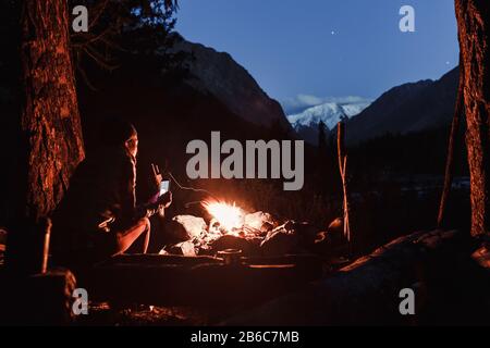 Femme randonneur se trouve près du feu de joie dans le camp et admire les montagnes sous le ciel étoilé Banque D'Images