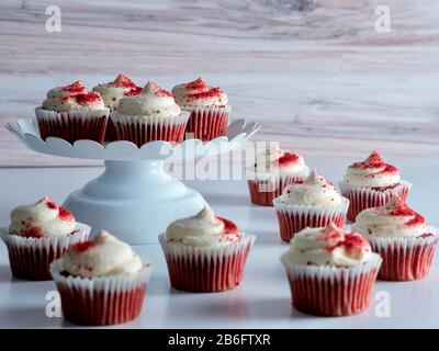 Une douzaine de petits cupcakes en velours rouge sur un plateau blanc et entourés d'autres sur la table en face. Délicieux dessert ! Banque D'Images