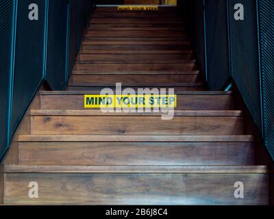 Attention À Votre pas, texte noir sur le panneau d'avertissement jaune sur l'escalier en bois. Escalier jusqu'à l'escalier avec panneau d'avertissement jaune. Banque D'Images