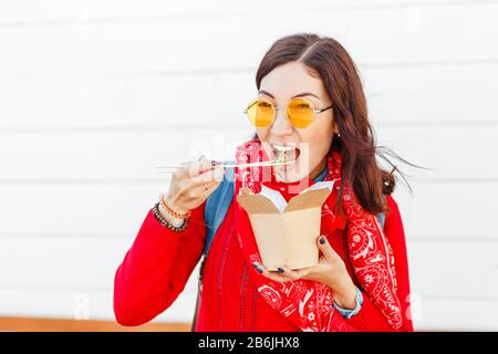 Femme hipster vif qui mange de la nourriture rapide asiatique à emporter avec des bâtonnets de hache, concept de nouilles wok Banque D'Images