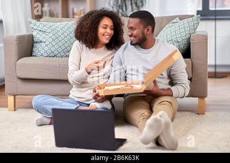 loisirs, gens et concept de la nourriture - couple afro-américain heureux avec ordinateur portable manger des pizzas à emporter à la maison Banque D'Images