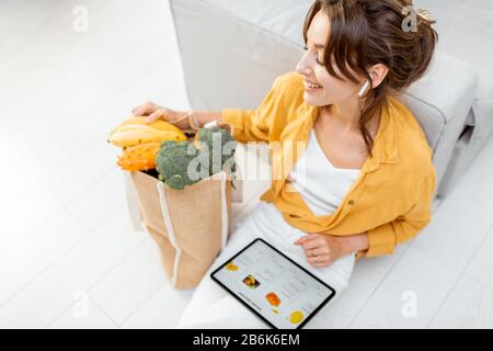 Jeune femme magasinage en ligne à l'aide d'une tablette numérique, assis avec sac plein de produits à la maison. Concept d'achat en ligne à l'aide d'appareils mobiles Banque D'Images