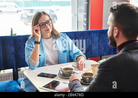 Jeune femme d'affaires séduisante qui ajuste les lunettes et discute avec un collègue lors d'une réunion dans un café moderne Banque D'Images