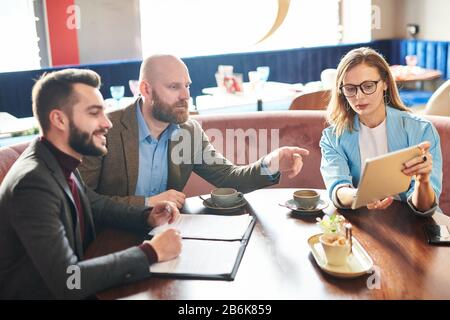 Femme d'affaires moderne dans des verres assis à la table et montrant des informations sur la tablette aux collègues pendant qu'ils discutent du contenu du contrat Banque D'Images