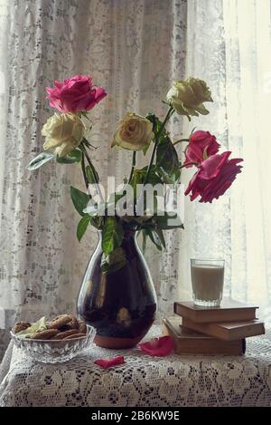 Un bouquet de roses dans un vase en céramique . Il y a à côté des livres, un verre de lait et un bol de cookies. Banque D'Images