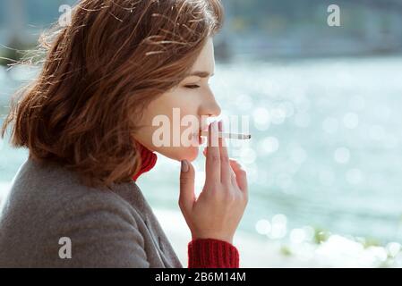 belle élégante triste stressante jeune femme en chandail et manteau fumer la cigarette près de la rivière Banque D'Images