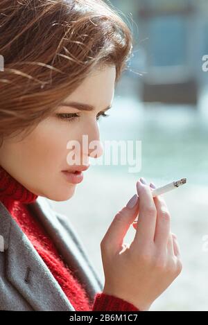 belle élégante triste stressante jeune femme en chandail et manteau fumer la cigarette près de la rivière Banque D'Images