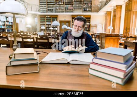 Littérature, classique, bibliothèque, concept d'éducation. Le lecteur d'homme senior se trouve dans l'intérieur de la bibliothèque ancienne de luxe et lit le livre. Homme barbu au visage heureux Banque D'Images