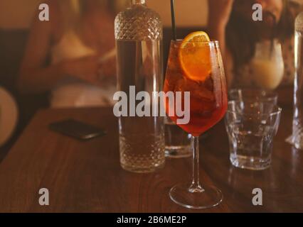 Wineglass avec boisson fraîche à l'alcool avec tranche orange et paille placée sur table en bois à côté de la bouteille en verre dans le bar avec des femmes floues dans le backgroun Banque D'Images