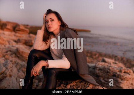 Une jeune femme pensive dans des vêtements élégants et chauds regardant l'appareil photo tout en étant assise sur la plage en automne soir Banque D'Images