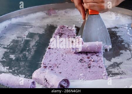 La préparation de la glace, de la cuisine thaïlandaise de rue le cuisinier tord habilement le violet congelé en rouleaux. Banque D'Images