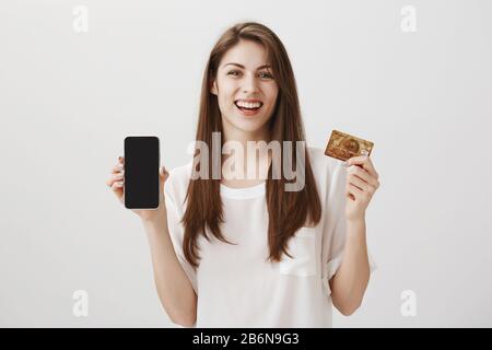 J'ai acheté un smartphone en crédit et j'en ai été fier. Photo studio d'une jeune étudiante émotive heureuse montrant une carte bancaire et un nouveau téléphone, se vanant des nouveautés Banque D'Images