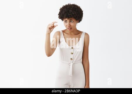 Mignon petite amie afro-américain perplexe avec la peau foncée et de l'afro hairstyle façonner à l'aide de petite taille, à la recherche tout en montrant la chose minuscule et Banque D'Images