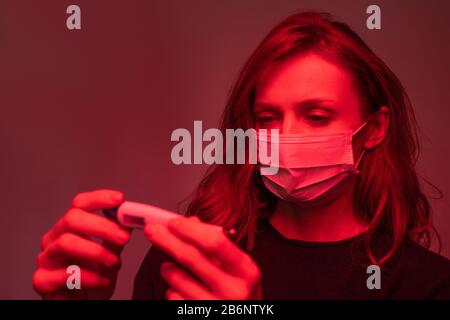 Jeune femme en masque de visage debout en lumière rouge et en vérifiant la température sur le thermomètre numérique Banque D'Images