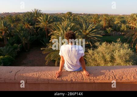 Désert marocain et palmiers avec un jeune modèle assis sur un mur d'argile Banque D'Images