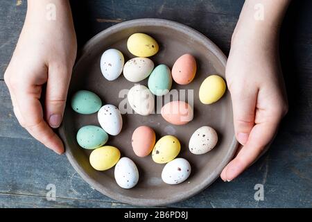 Les mains tenant des œufs de pâques peints dans des tons pastel élégants dans une plaque en céramique grise sur fond vieux bois vintage, Joyeuses Pâques Banque D'Images