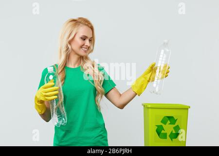 Une jeune femme bénévole dans des tee-shirts et des gants verts collecte et jette les déchets plastiques dans un récipient pour recyclage. Fond de studio blanc Banque D'Images