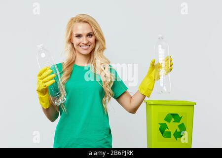 Une jeune femme bénévole dans des tee-shirts et des gants verts collecte et jette les déchets plastiques dans un récipient pour recyclage. Fond de studio blanc Banque D'Images