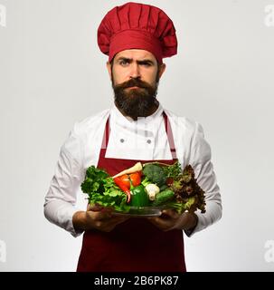 Faire cuire avec un visage sérieux en bordeaux en uniforme contient des ingrédients de salade. Concept de cuisine et de régime végétarien. Le chef tient un bol avec des légumes frais. Homme avec barbe isolé sur fond blanc. Banque D'Images