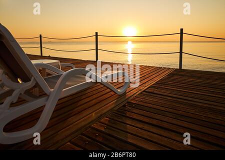Le Beach-transat se tient sur une jetée en bois sur le fond d'une mer calme et du soleil levant. Banque D'Images