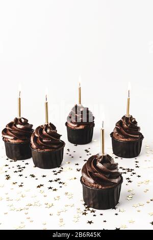 Délicieux cupcakes au chocolat avec glaçage au chocolat, paillettes d'or sautées et bougies brûlantes sur fond blanc avec confetti en forme d'étoile dorée. H Banque D'Images