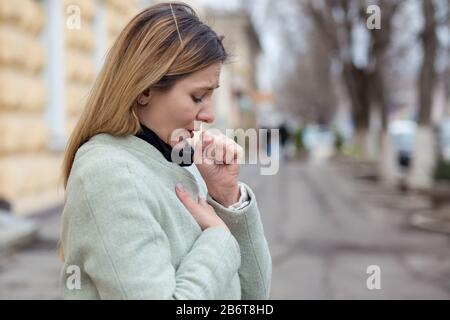 Une jeune fille malade, une jeune femme blonde toussant le sentiment mauvais, malade, étant frappé par le coronavirus, Covid2019 debout à l'extérieur dans la ville. Banque D'Images