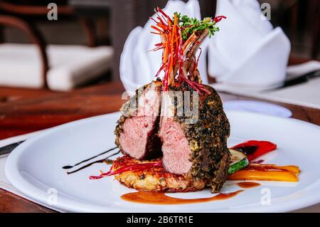 Bifteck rôti d'agneau avec croûte aux herbes croustillantes et légumes grillés sur la plaque blanche à la table du dîner Banque D'Images