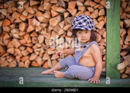 Un beau petit enfant dans une combinaison grise et un capuchon dans une cage se trouve près d'un mur de bois de chauffage, regarde l'appareil photo. Banque D'Images