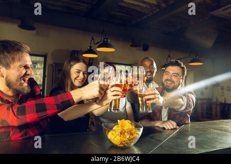 Les amateurs de sport applaudisent au bar, au pub. Prendre des verres à bière tout en regardant le championnat, la compétition. Groupe multiethnique d'amis enthousiasmés par la traduction. Émotions humaines, expression, concept de soutien. Banque D'Images