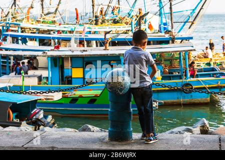 Jeune garçon se tenant sur le port en regardant les pêcheurs sur leurs bateaux de pêche, Sandakan, Sabah district, Bornéo, Malaisie, Asie Banque D'Images