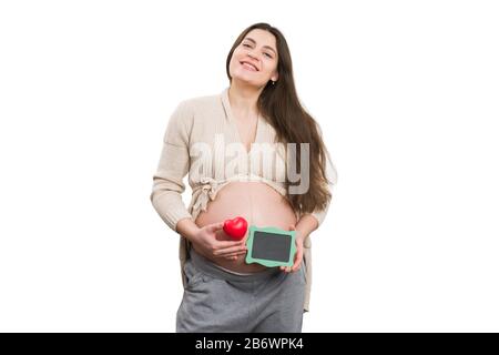 Femme enceinte souriante tenant le jouet de coeur et le blackboard vert isolé sur fond blanc Banque D'Images