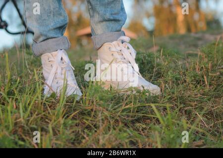 Gros plan sur les pieds de l'adolescent dans des sneakers blanches modernes et tendance et des jeans roulés. Jour d'été ensoleillé, fond d'herbe verte. Banque D'Images