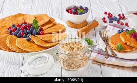 gros plan de crêpes au gruau servies avec des bleuets frais et des canneberges sur une plaque blanche avec des ingrédients, une fourchette dorée et un couteau, une sauce aux canneberges Banque D'Images