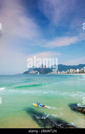 Un surfeur masculin entrant dans l'océan Atlantique aux roches d'Arpoador avec la plage d'Ipanema, le quartier et les montagnes de Dois Irmaos en arrière-plan Banque D'Images