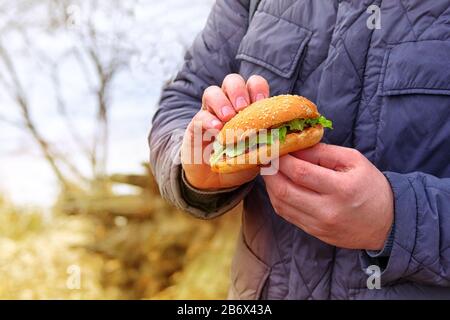 Hamburger avec un pain blanc aux graines de sésame. Pause déjeuner en voyage. Restauration rapide pour le déjeuner, à emporter. La main tient un délicieux hamburger frais. Banque D'Images