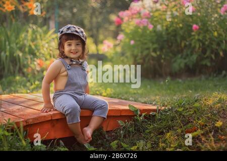 Joli petit garçon souriant assis sur un petit pont en bois dans un jardin fleuri en été. Journée ensoleillée dans le jardin. Banque D'Images