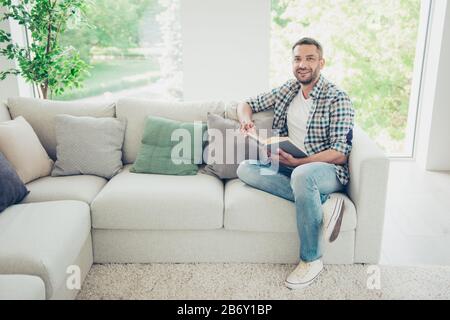 Portrait de beau homme de charme expert bibliothèque de sit divan ont des études inspirées se sentir gai contenu tenir main barbu plaid moderne denim jeans grand Banque D'Images