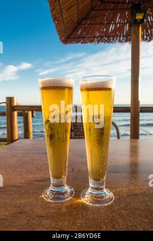 Deux verres de bière sur une table servis dans un restaurant de plage, avec mer méditerranéenne derrière. Banque D'Images