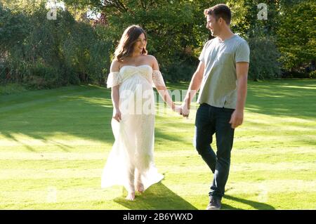 Belle jeune femme enceinte, portant une longue robe blanche, marchant main dans la main avec son partenaire lors d'une belle journée d'été Banque D'Images