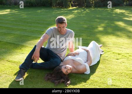 Belle jeune femme enceinte, portant une longue robe blanche, posée sur l'herbe avec son partenaire assis à côté d'elle Banque D'Images