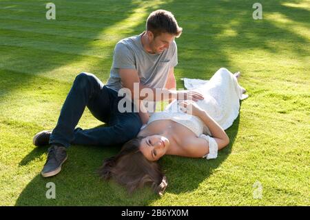 Belle jeune femme enceinte, portant une longue robe blanche, posée sur l'herbe avec son partenaire assis à côté d'elle Banque D'Images